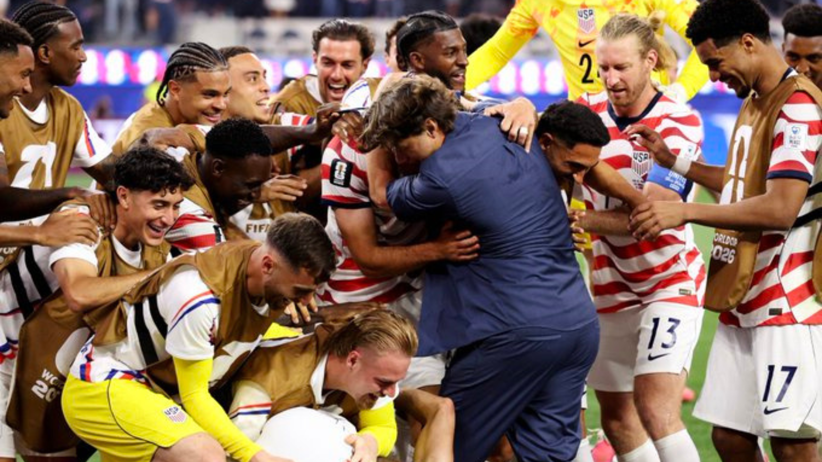 Soccer team in red-and-white striped jerseys celebrate, hugging and lifting a teammate on the field.