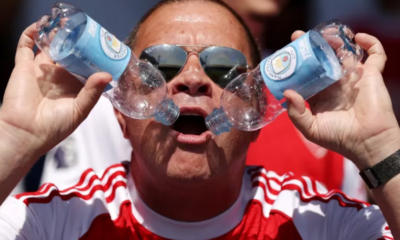Man wearing sunglasses and a red-and-white sports shirt holds two plastic water bottles pressed together in front of his open mouth, outdoors.