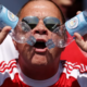 Man wearing sunglasses and a red-and-white sports shirt holds two plastic water bottles pressed together in front of his open mouth, outdoors.