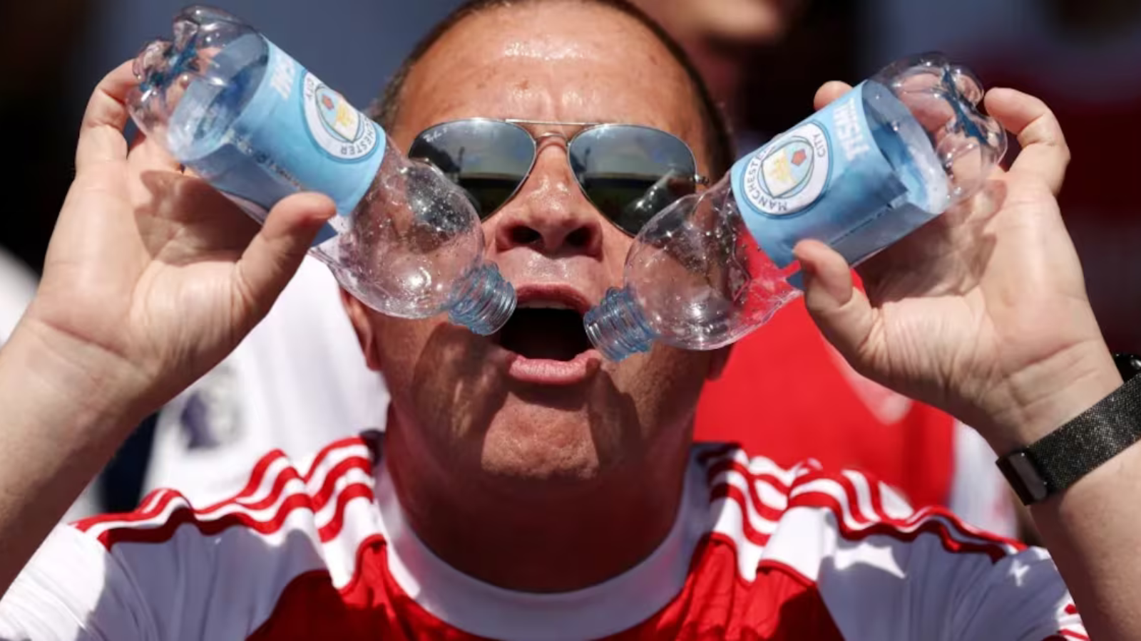 Man wearing sunglasses and a red-and-white sports shirt holds two plastic water bottles pressed together in front of his open mouth, outdoors.