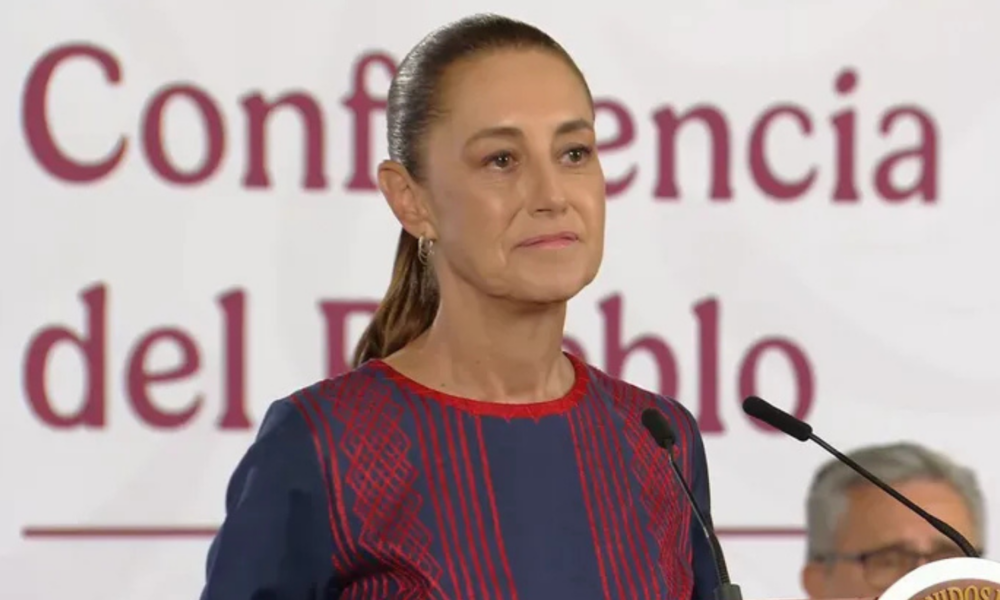 Woman stands at a podium giving a speech, wearing a navy dress with red embroidery and a high ponytail, microphones in front, banner behind she speaks at a formal event.