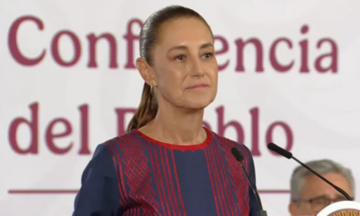 Woman stands at a podium giving a speech, wearing a navy dress with red embroidery and a high ponytail, microphones in front, banner behind she speaks at a formal event.
