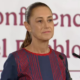 Woman stands at a podium giving a speech, wearing a navy dress with red embroidery and a high ponytail, microphones in front, banner behind she speaks at a formal event.