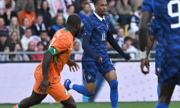 Soccer players in orange and blue kits sprint on the field as a defender closes in near a goal area, crowd watching in the stands behind.