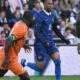 Soccer players in orange and blue kits sprint on the field as a defender closes in near a goal area, crowd watching in the stands behind.
