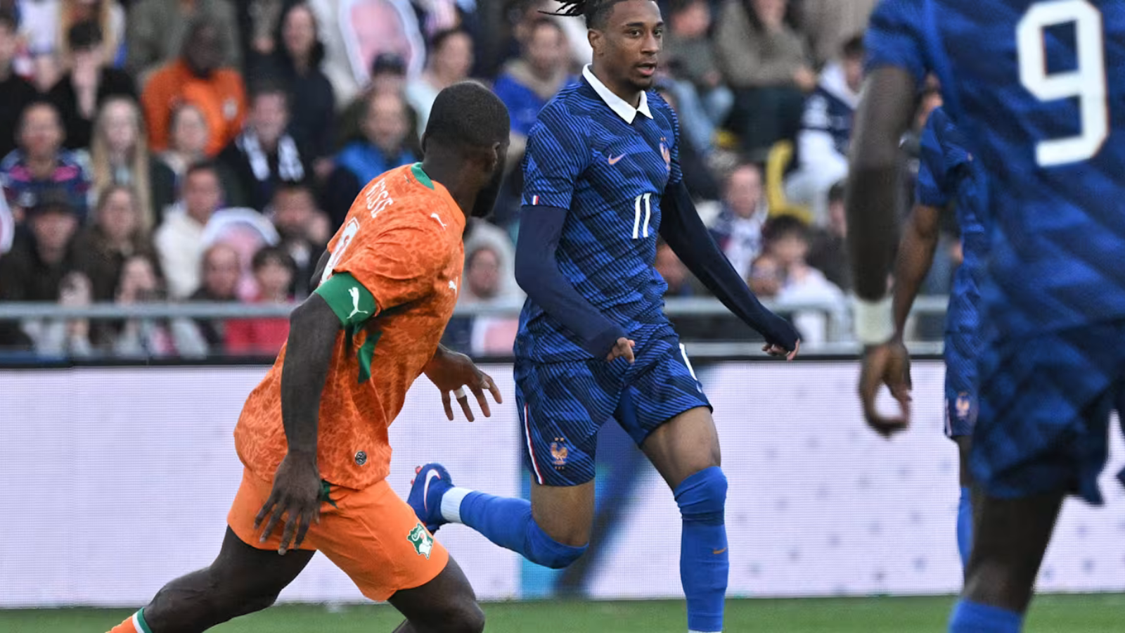 Soccer players in orange and blue kits sprint on the field as a defender closes in near a goal area, crowd watching in the stands behind.