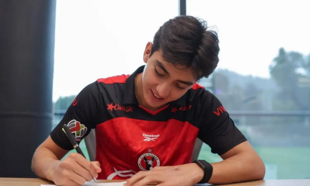 Young man in a red and black sports jersey signs a document at a table indoors, focused on the task.