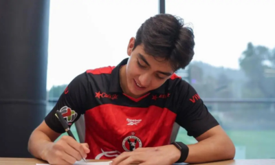 Young man in a red and black sports jersey signs a document at a table indoors, focused on the task.