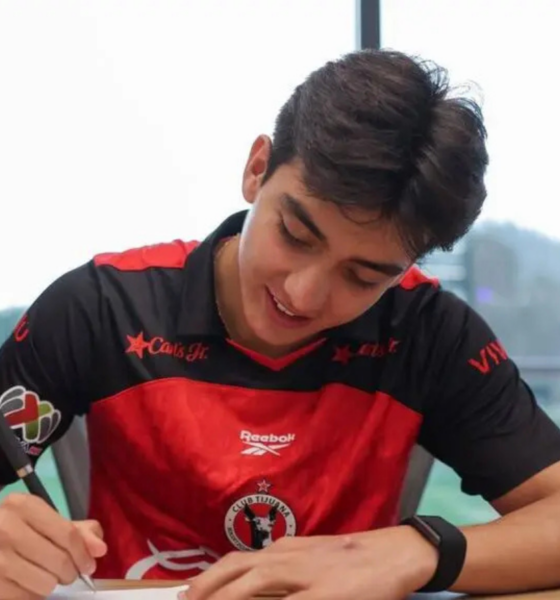 Young man in a red and black sports jersey signs a document at a table indoors, focused on the task.
