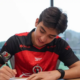 Young man in a red and black sports jersey signs a document at a table indoors, focused on the task.