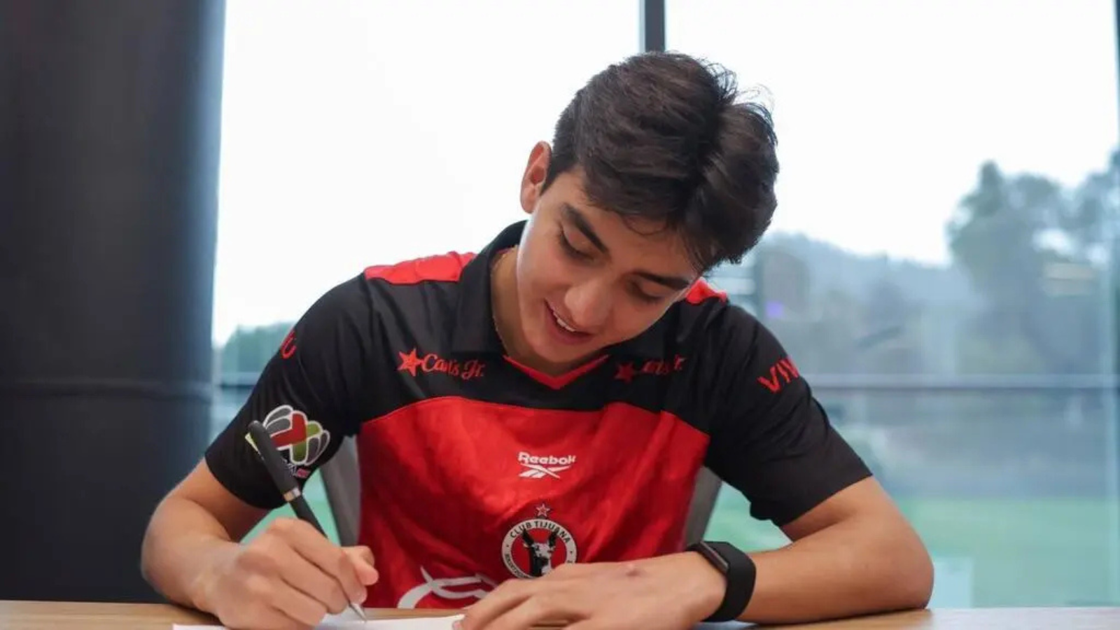 Young man in a red and black sports jersey signs a document at a table indoors, focused on the task.