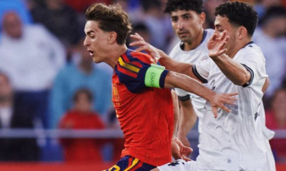Soccer player in a red kit with a green captain's armband sprinting forward as two white-jersey opponents pursue him.