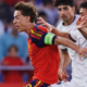 Soccer player in a red kit with a green captain's armband sprinting forward as two white-jersey opponents pursue him.