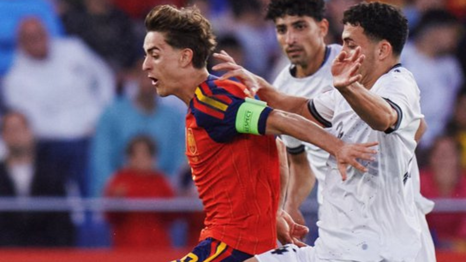 Soccer player in a red kit with a green captain's armband sprinting forward as two white-jersey opponents pursue him.