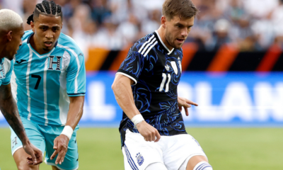 Soccer game action: a player in a dark blue patterned kit (11) dribbles the ball while two teal-clad opponents close in.