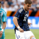 Soccer game action: a player in a dark blue patterned kit (11) dribbles the ball while two teal-clad opponents close in.