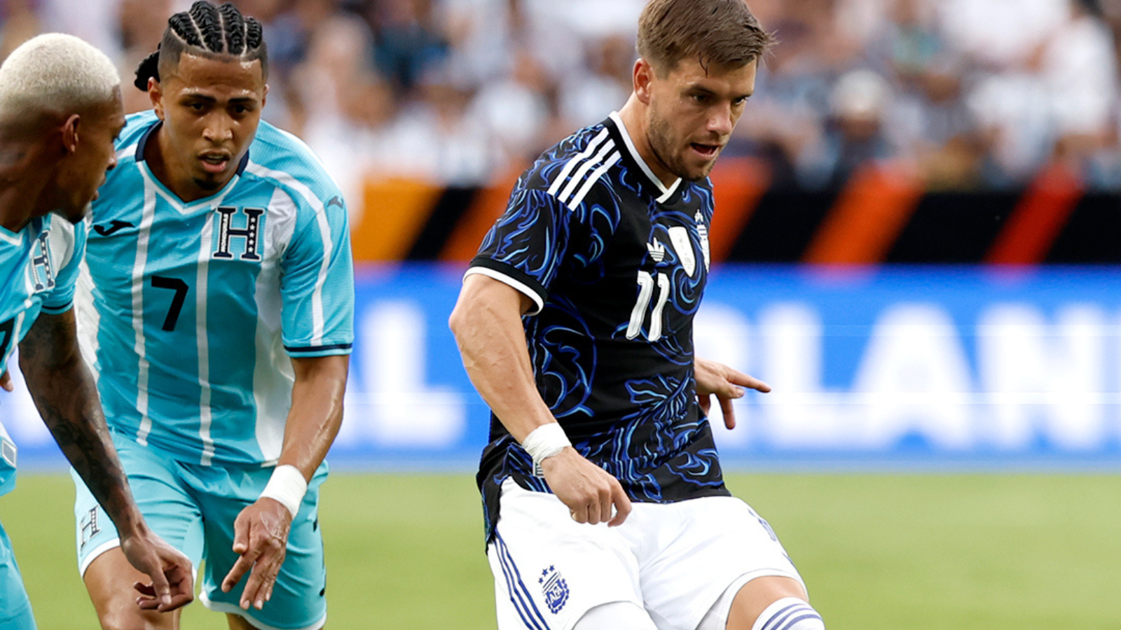 Soccer game action: a player in a dark blue patterned kit (11) dribbles the ball while two teal-clad opponents close in.