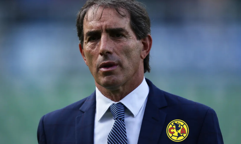 Man in a dark blue suit and white shirt with a blue patterned tie, standing outdoors; wearing Club América badge on his chest.
