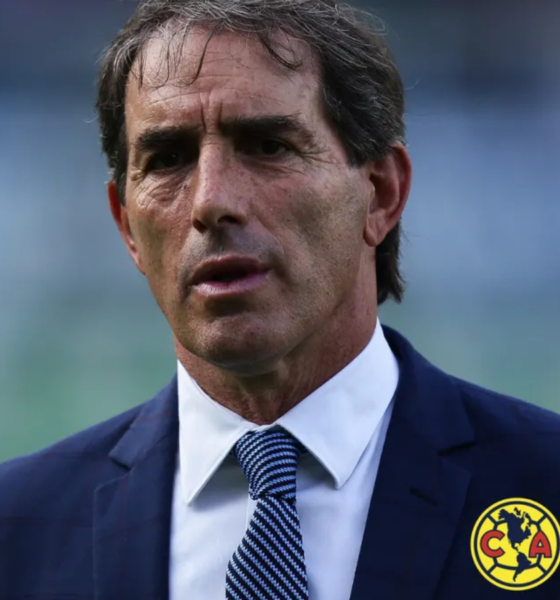 Man in a dark blue suit and white shirt with a blue patterned tie, standing outdoors; wearing Club América badge on his chest.