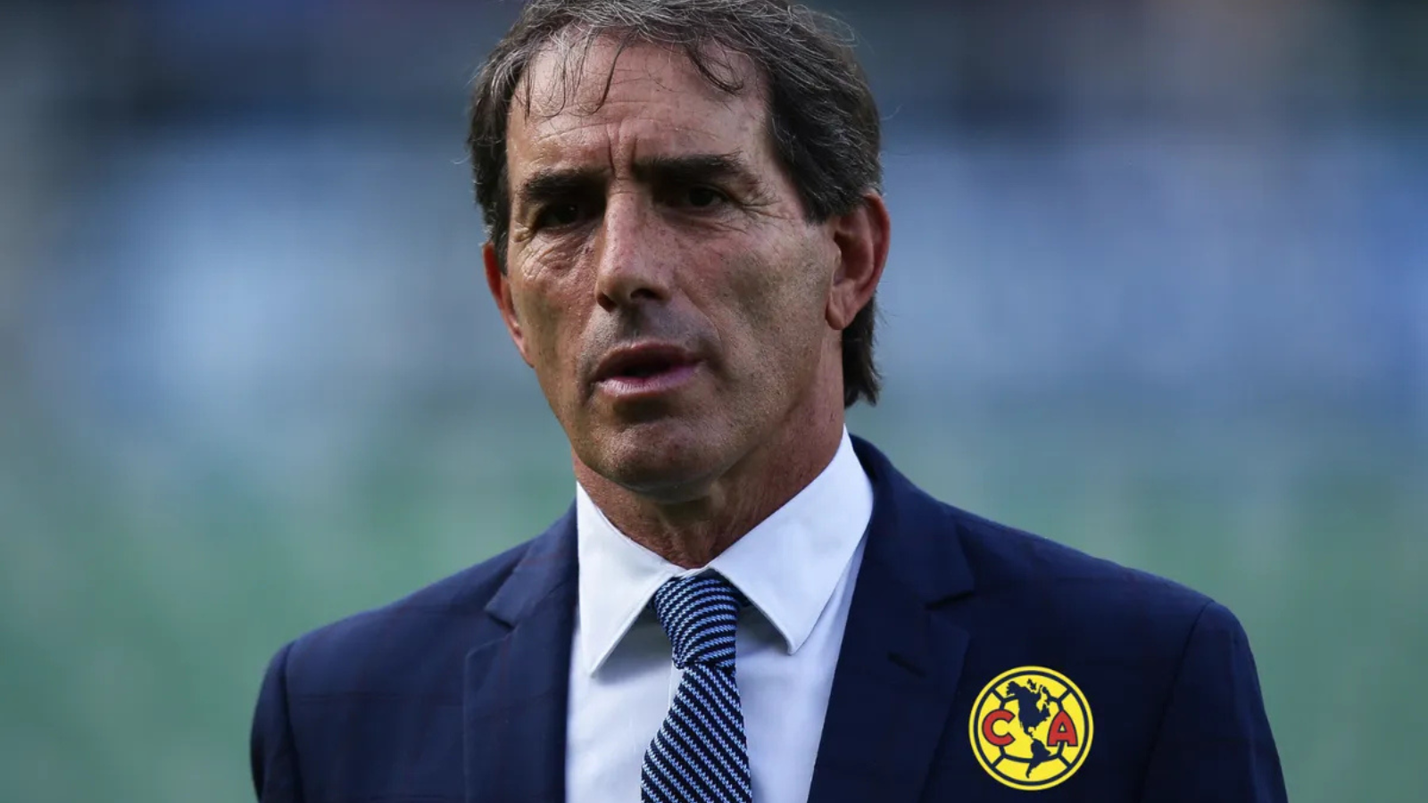 Man in a dark blue suit and white shirt with a blue patterned tie, standing outdoors; wearing Club América badge on his chest.