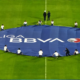 A large blue banner reading 'LIGA BBVA' laid out on a green soccer field with people around it