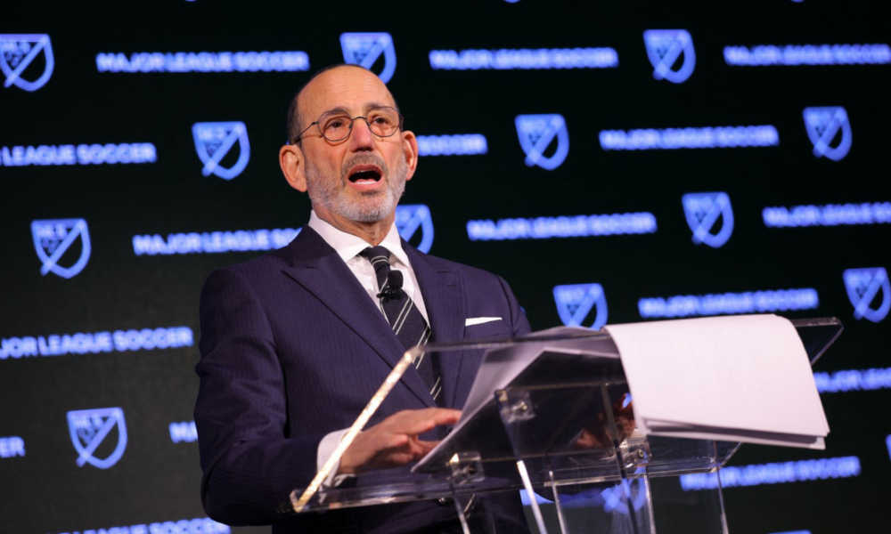Man in a dark pinstripe suit and glasses speaks at a clear podium with MLS branding in the background, delivering a formal address.