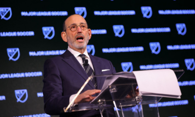 Man in a dark pinstripe suit and glasses speaks at a clear podium with MLS branding in the background, delivering a formal address.