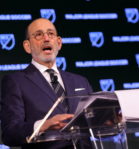 Man in a dark pinstripe suit and glasses speaks at a clear podium with MLS branding in the background, delivering a formal address.