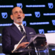 Man in a dark pinstripe suit and glasses speaks at a clear podium with MLS branding in the background, delivering a formal address.