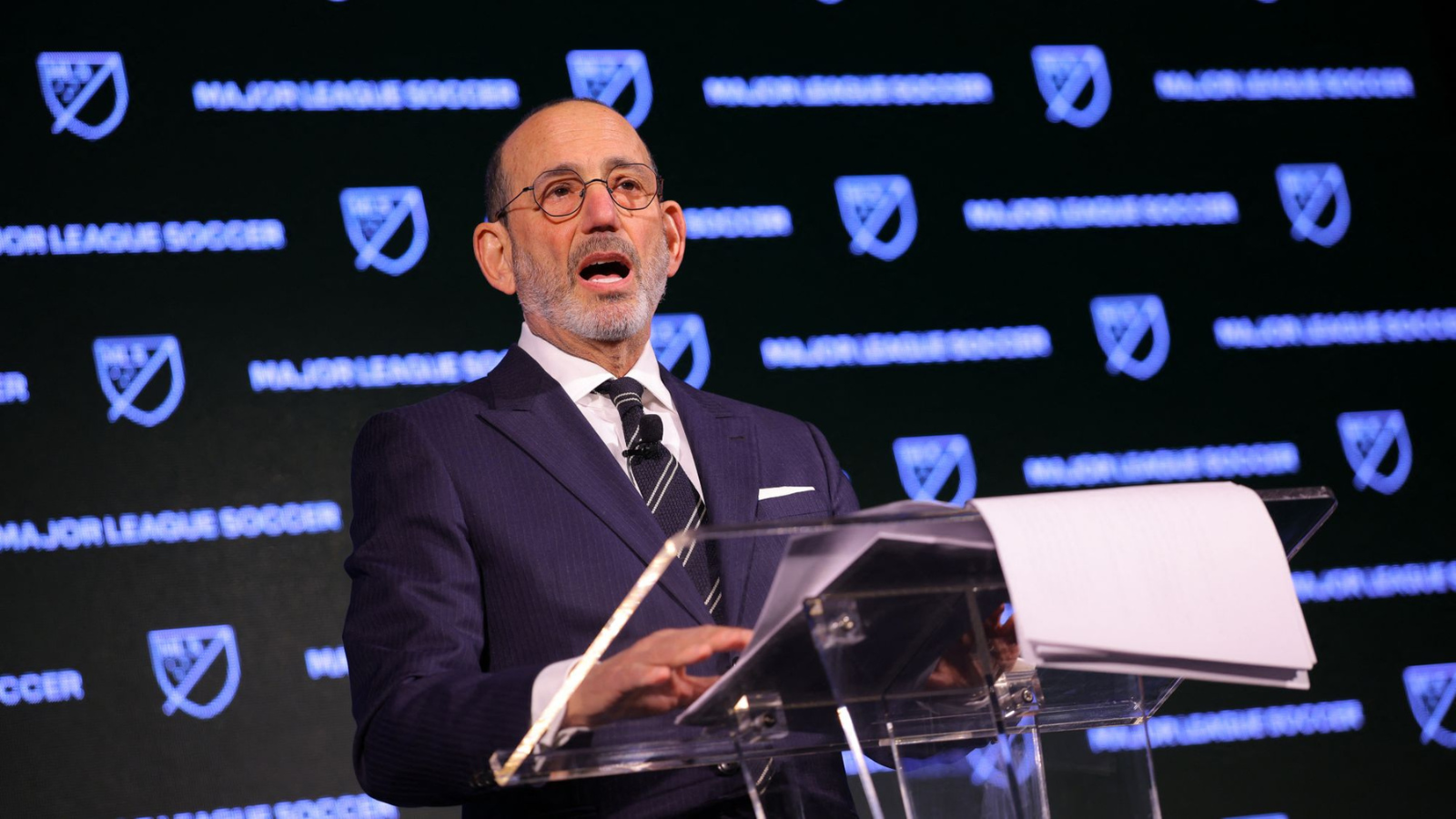 Man in a dark pinstripe suit and glasses speaks at a clear podium with MLS branding in the background, delivering a formal address.