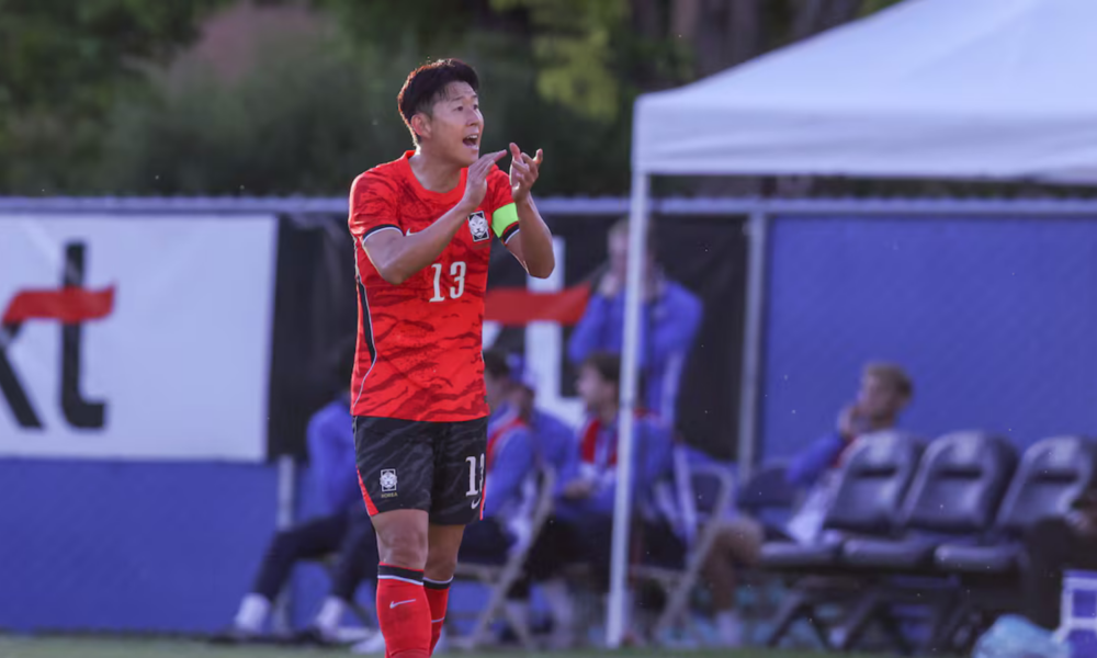 Soccer player wearing red jersey #13 on the field, clapping with teammates on the sidelines in the background.