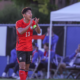Soccer player wearing red jersey #13 on the field, clapping with teammates on the sidelines in the background.