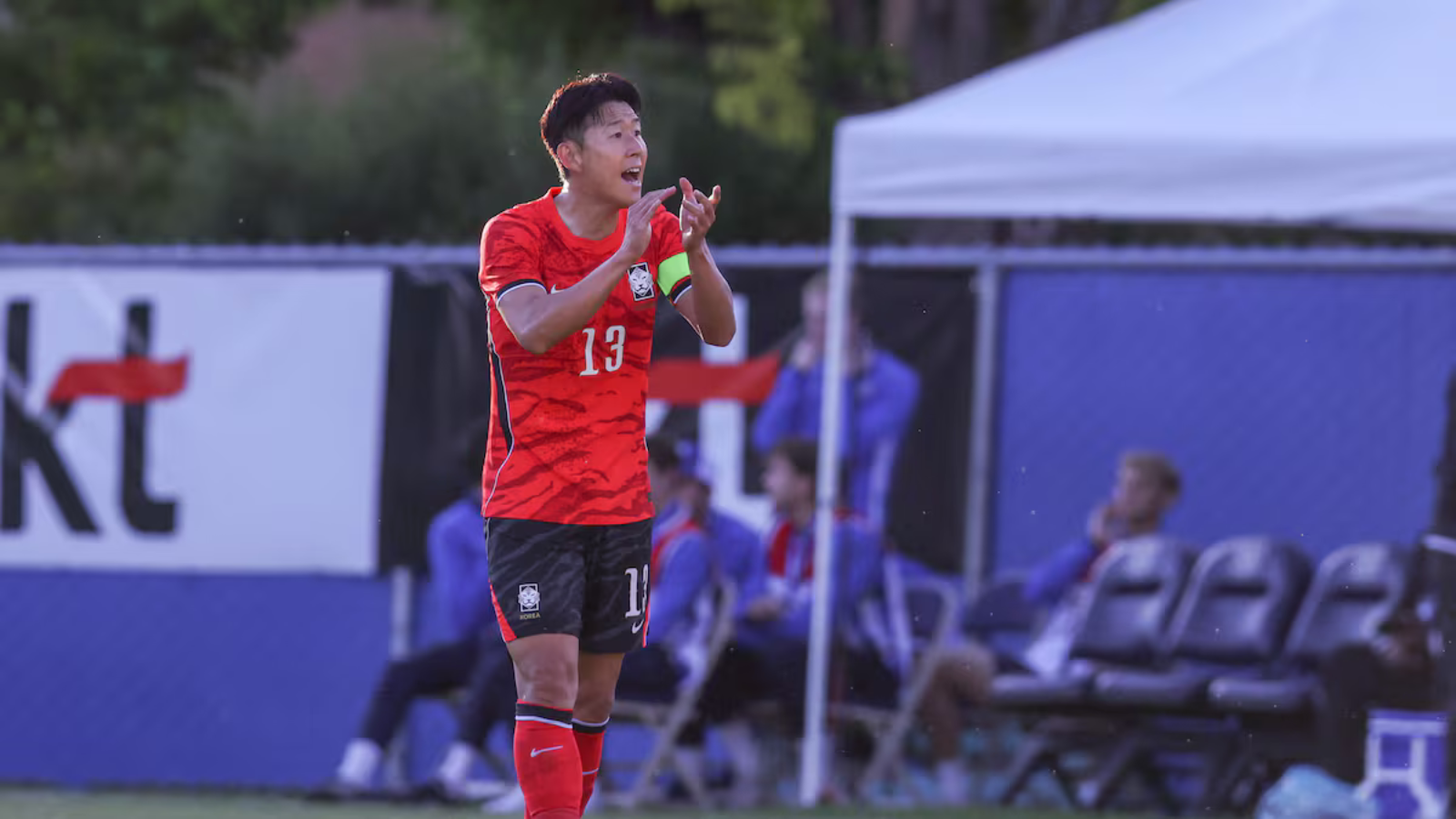 Soccer player wearing red jersey #13 on the field, clapping with teammates on the sidelines in the background.