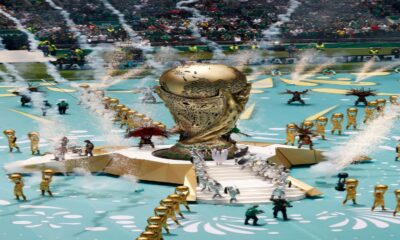 Giant golden trophy centerpiece on a turquoise parade stage with dancers in silver and gold, fountains spraying water, and a cheering stadium crowd in the background.