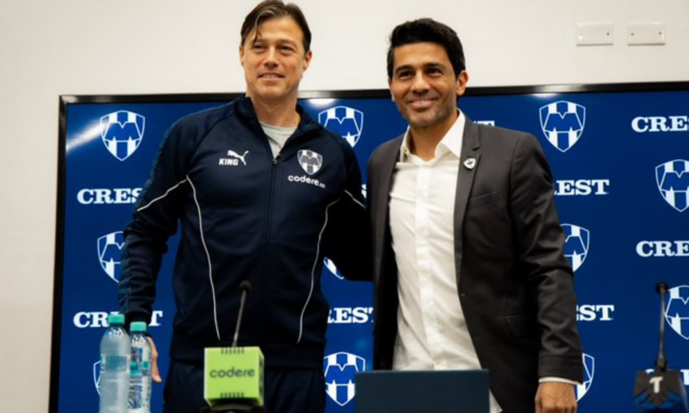 Two men stand together at a press conference, one in a navy tracksuit and the other in a suit, smiling in front of a blue Crest backdrop with logos.