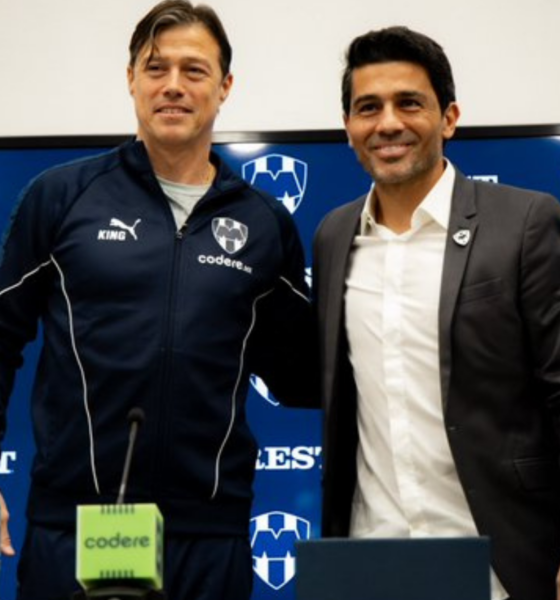 Two men stand together at a press conference, one in a navy tracksuit and the other in a suit, smiling in front of a blue Crest backdrop with logos.