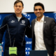 Two men stand together at a press conference, one in a navy tracksuit and the other in a suit, smiling in front of a blue Crest backdrop with logos.