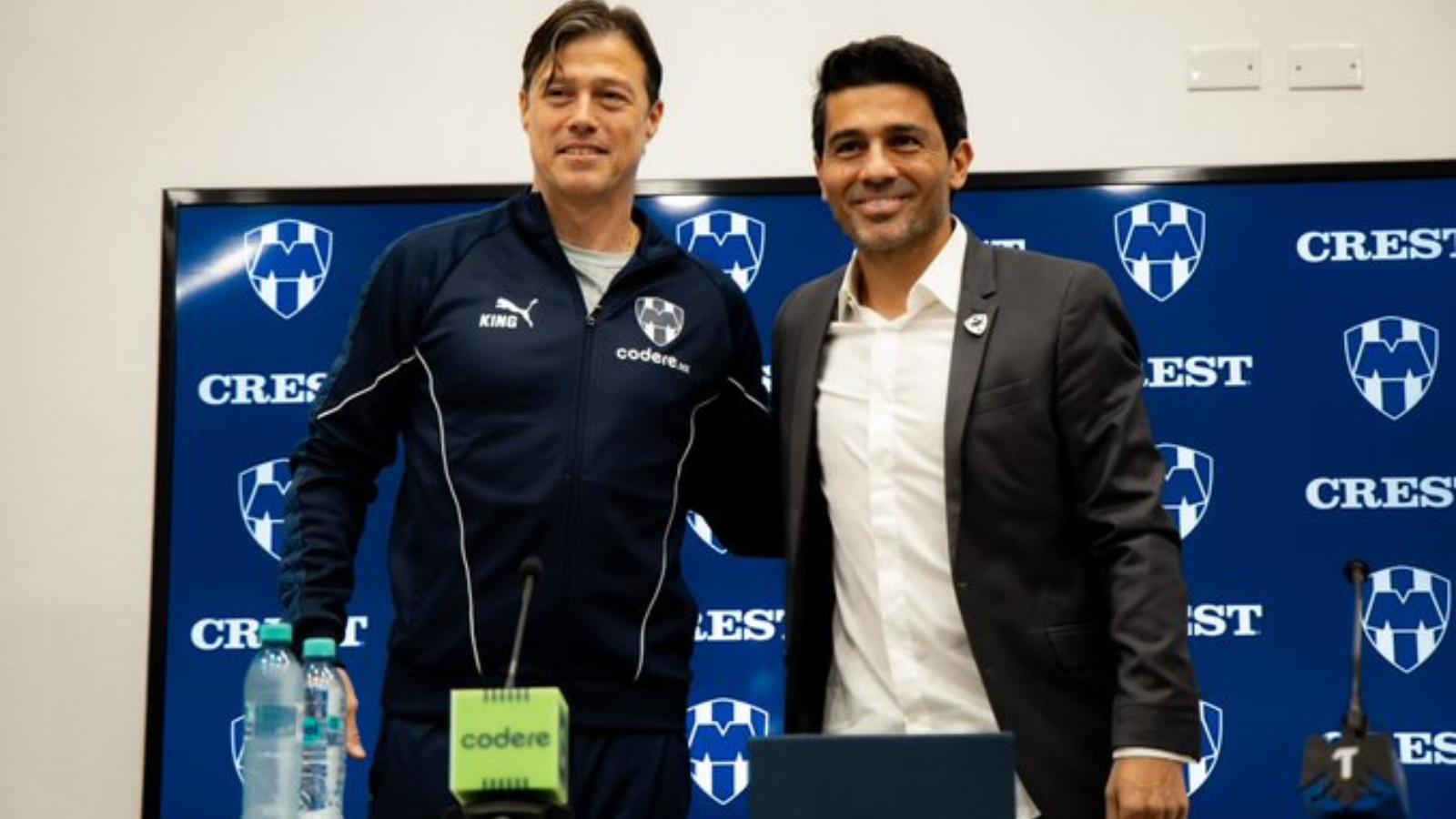 Two men stand together at a press conference, one in a navy tracksuit and the other in a suit, smiling in front of a blue Crest backdrop with logos.