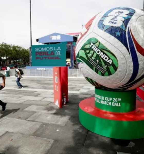 Giant soccer ball sculpture on a green-red pedestal in a plaza, with a banner reading 'Somos la Perla del Futbol' in the background.