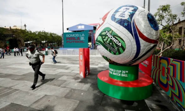 Giant soccer ball sculpture on a green-red pedestal in a plaza, with a banner reading 'Somos la Perla del Futbol' in the background.