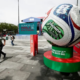 Giant soccer ball sculpture on a green-red pedestal in a plaza, with a banner reading 'Somos la Perla del Futbol' in the background.