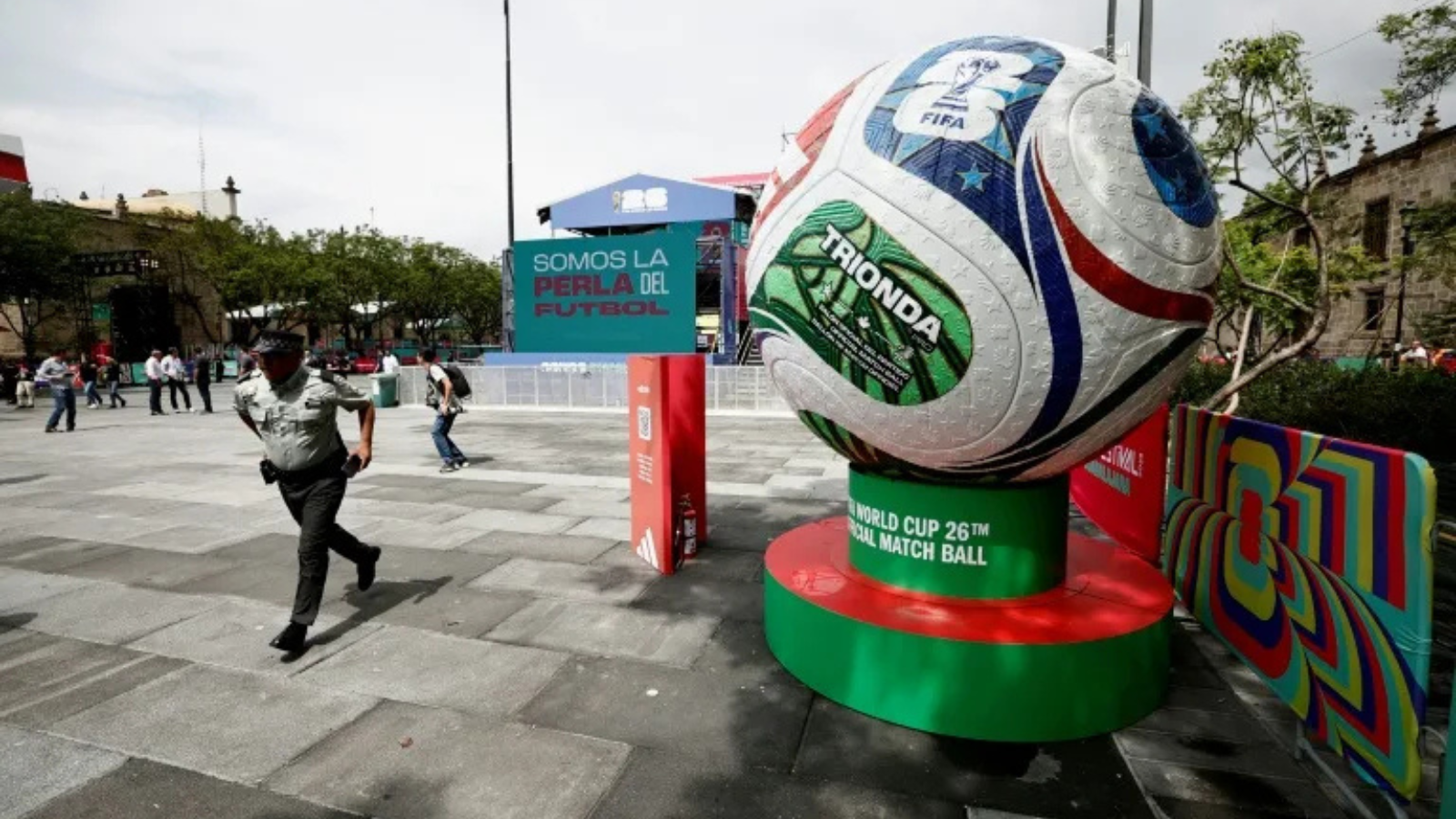 Giant soccer ball sculpture on a green-red pedestal in a plaza, with a banner reading 'Somos la Perla del Futbol' in the background.