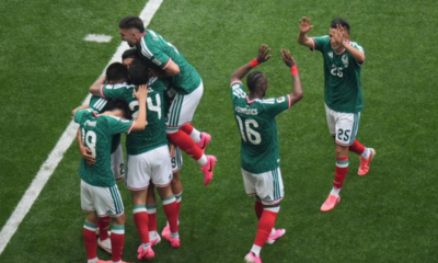Group of soccer players in green jerseys celebrate together on a grass pitch, embracing and jumping in celebration after a goal