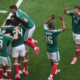 Group of soccer players in green jerseys celebrate together on a grass pitch, embracing and jumping in celebration after a goal