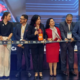Group of professionals in formal attire cutting a ribbon at a ceremony on a stage with a blue backdrop.