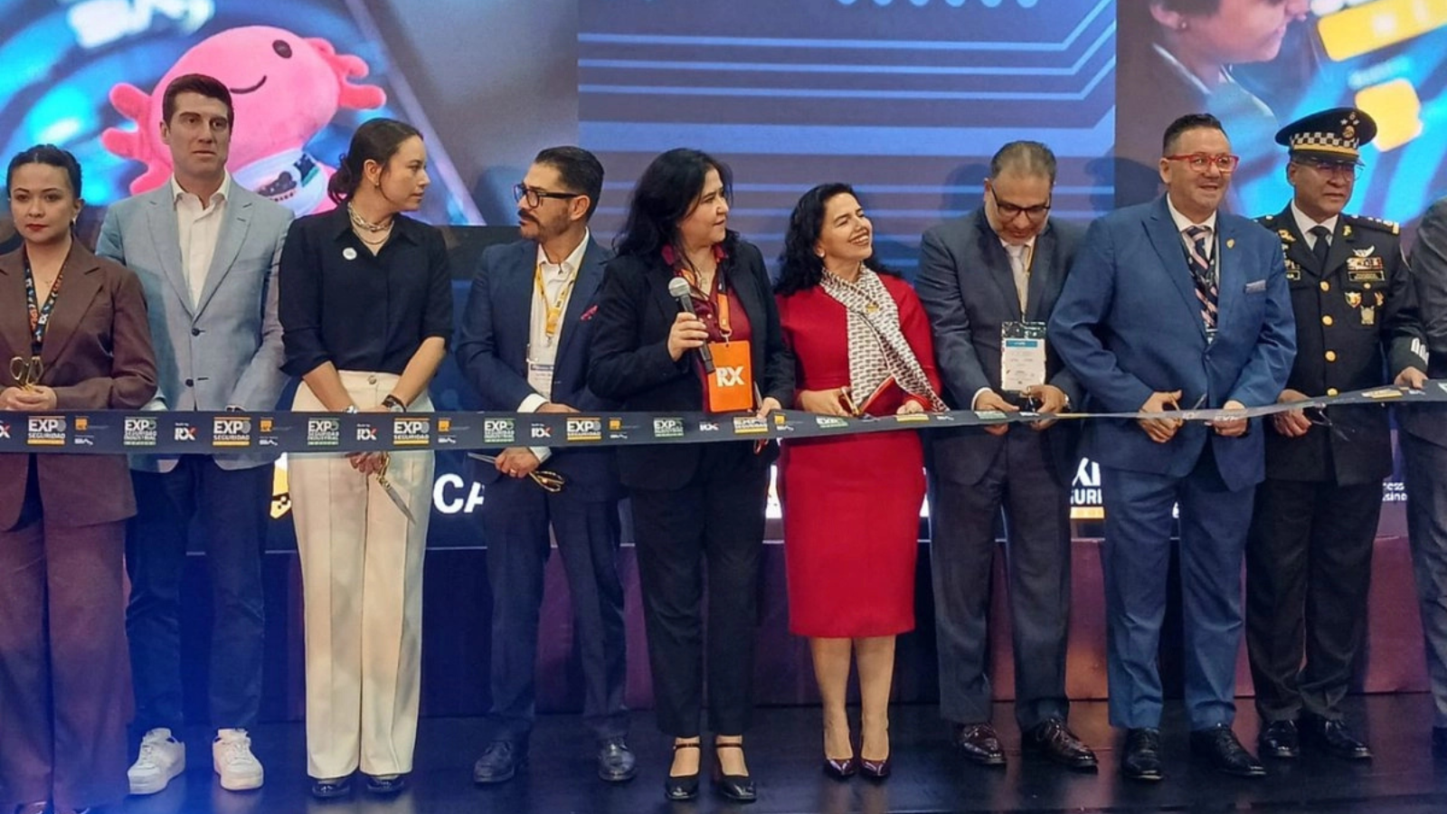 Group of professionals in formal attire cutting a ribbon at a ceremony on a stage with a blue backdrop.