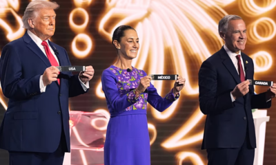Three adults on a brightly lit stage hold black cards labeled USA, MEXICO, and CANADA in front of a swirling golden backdrop.