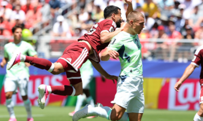 Soccer players in maroon and light-green kits clash for the ball during a sunny match; one player leaps while another runs ahead on the field.