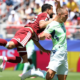 Soccer players in maroon and light-green kits clash for the ball during a sunny match; one player leaps while another runs ahead on the field.