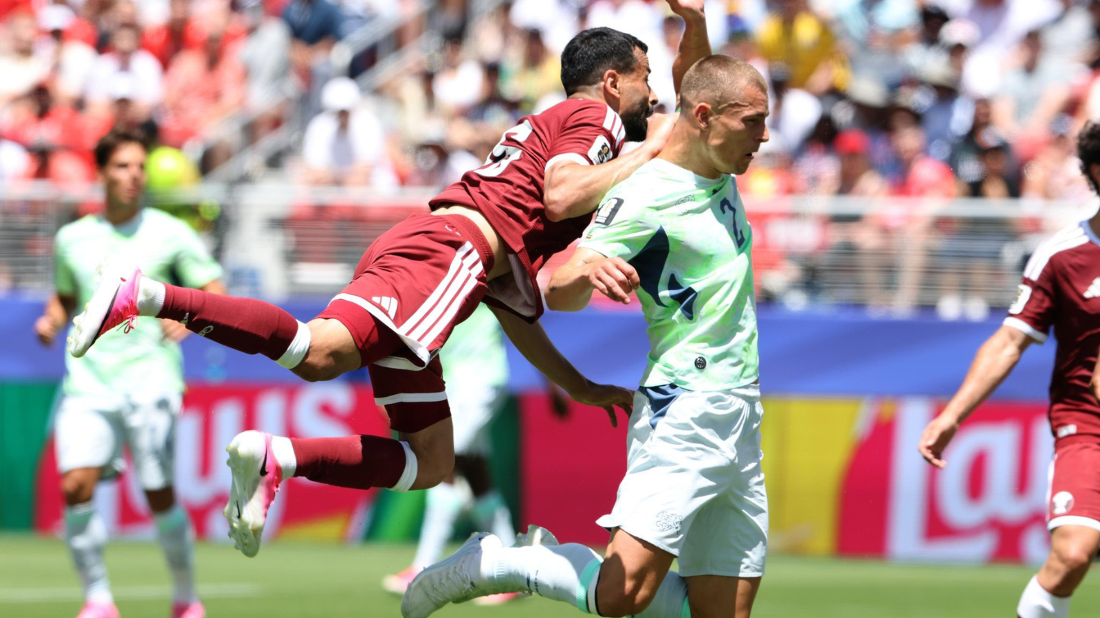 Soccer players in maroon and light-green kits clash for the ball during a sunny match; one player leaps while another runs ahead on the field.